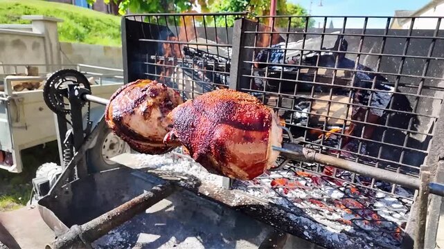 Whole pork shanks roasting slowly on a rotating spit outdoors, over glowing coals. Large pieces of pork rotate slowly on a metal spit over glowing embers and ashes, giving off a delicious aroma. 