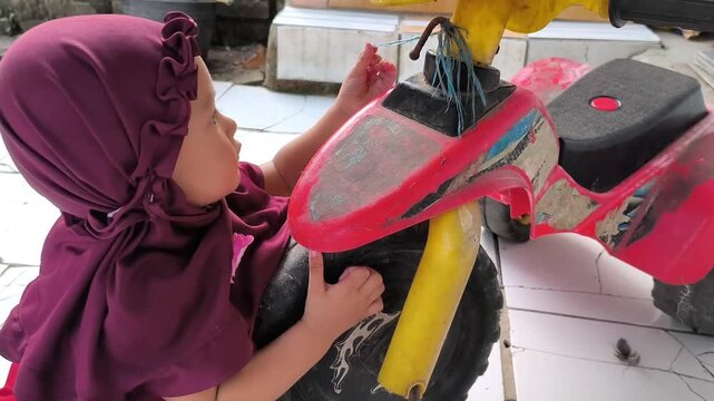 Video footage of a Muslim toddler girl playing with a red plastic tricycle at home.