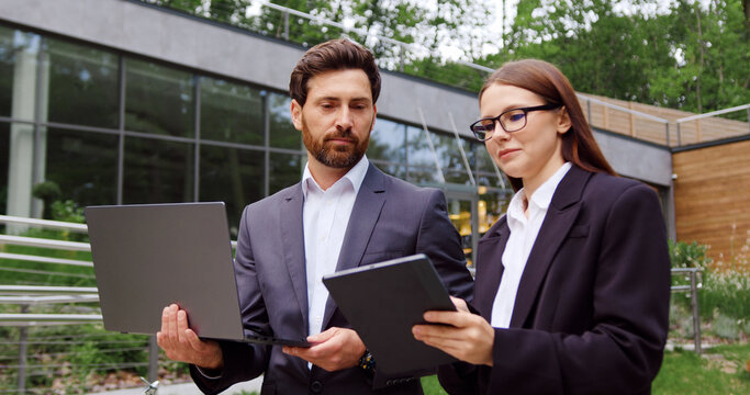 Businessman and businesswoman walk to the office while having a business conversation and looking at a tablet held by the woman, with the man carrying a laptop.