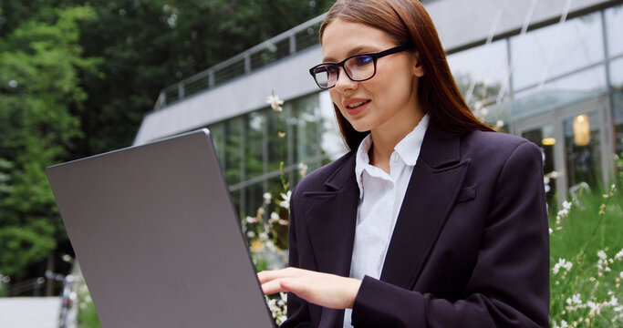 Young businesswoman in formal attire typing on a laptop outdoors in a city park, with a modern glass office building in the background. Concept of business, remote work, productivity, career