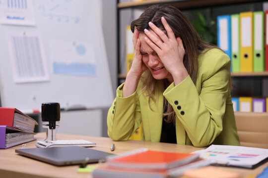 Young woman in lime blazer sits at desk covering face with hands during stressful work moment inside busy office, surrounded by laptop, papers, folders, whiteboard and binders, with stressed, stress