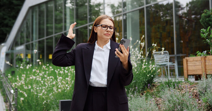 Young Caucasian woman walking in a city park, looking disappointed and concerned as she reads an unexpected message on her smartphone.