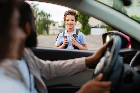Smiling black schoolboy with backpack standing outside the car looking at parents