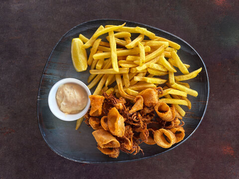 A delicious plate of fried calamari with French fries, served with a small bowl of sauce and a lemon wedge on a dark plate.