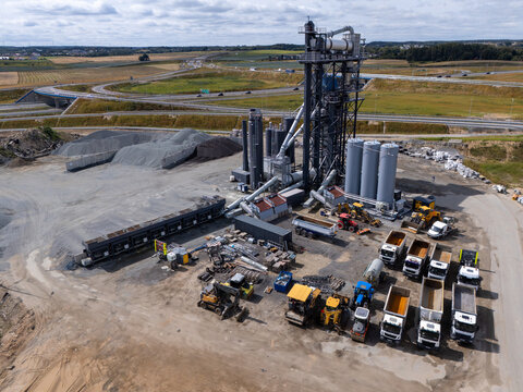 Aerial view of an asphalt plant featuring storage silos, heavy construction machinery, and parked trucks near a highway in Gdansk, Poland.