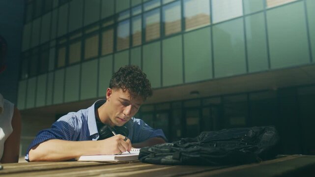 A focused high school student is diligently writing notes in a notebook at an outdoor table. He is engaged in study, perhaps on a university or school campus on a bright day.