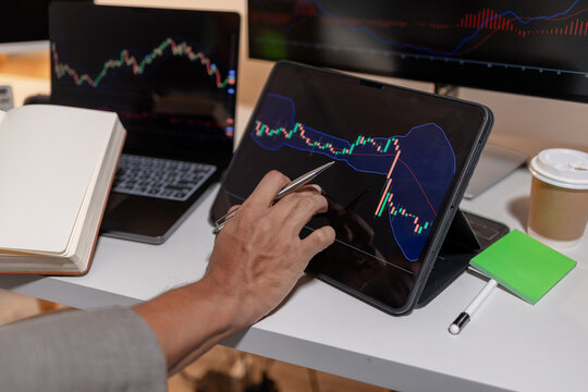 Professional trader analyzing stock market charts across multiple screens using laptop and tablet to monitor price movements, technical indicators and investment strategies in modern trading workspace