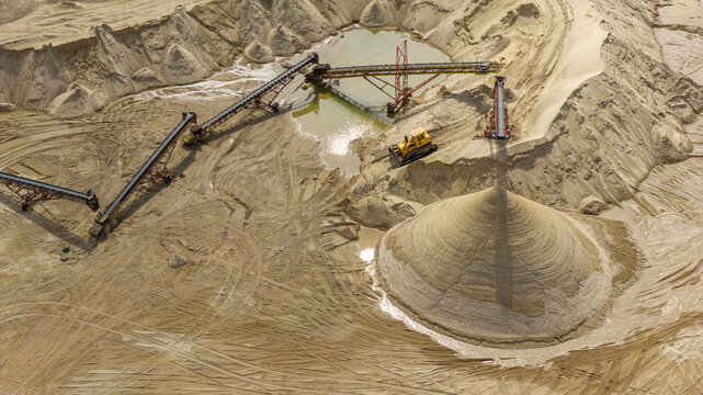 Aerial view of a sand quarry featuring conveyor belts, a bulldozer, a large pile of sand, and a small pool of water in Poland.