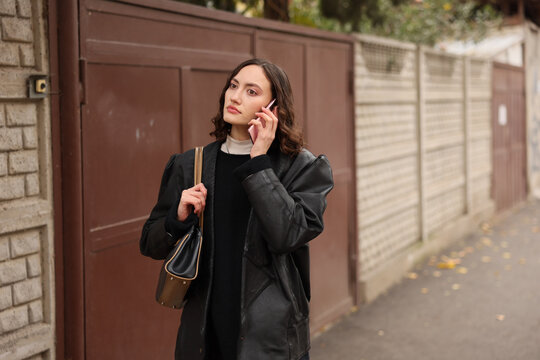 Young woman in black outerwear talks on smartphone beside residential gate, carrying shoulder bag and looking off camera with calm thoughtful mood on quiet city sidewalk in soft daylight