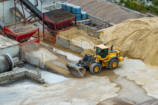Aerial view of a yellow wheel loader operating in a cement production plant with piles of sand, gravel, and industrial conveyor equipment in Poland.