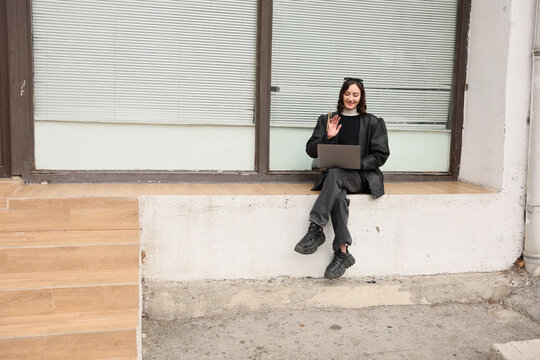 Young woman sits outdoors on low ledge with laptop on her lap and smiles while waving, wearing dark casual clothes beside large window with blinds in calm urban setting, remote work concept