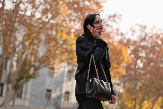 Young woman in black coat talks on smartphone while walking through autumn city street, carrying shoulder bag and looking away in soft warm light with blurred trees and building backdrop