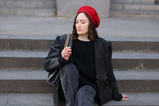 Young woman rests on concrete steps in black coat, dark sweater, and dark jeans, holding cigarette with calm pensive mood in muted urban setting, showing relaxed street fashion style