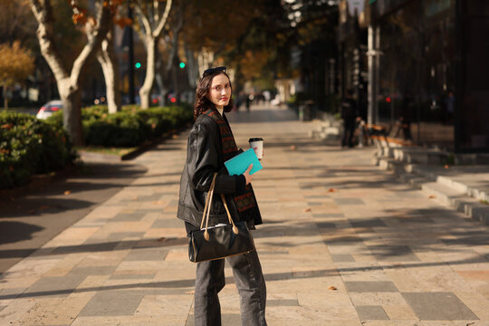 Young woman in dark outfit stands on sunlit urban sidewalk holding coffee cup, turquoise notebook, and handbag, looking toward camera with calm expression, autumn trees and softly blurred street