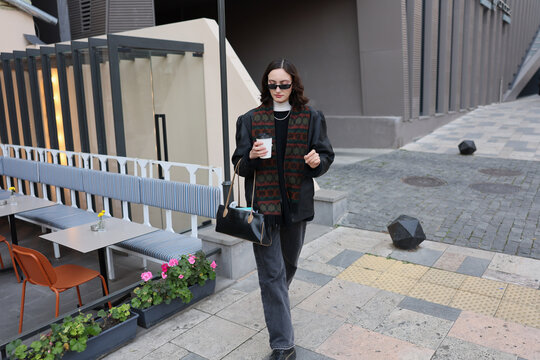 Stylish young woman walks through modern city caf terrace with takeaway coffee cup and handbag, wearing sunglasses, dark clothing, and patterned scarf beside outdoor seating and contemporary