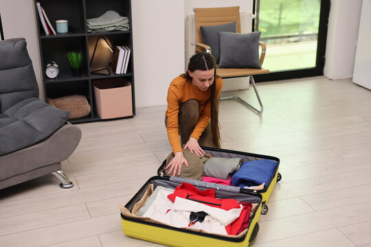 Young woman kneels beside open suitcase while packing folded clothes in bright modern living room, with organized shelves, cushioned chair, light wood floor, and daylight from large window