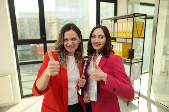 Two confident women smile and give thumbs up in bright modern office, holding folders and documents near large windows, shelves of binders, warm daylight, corporate success atmosphere