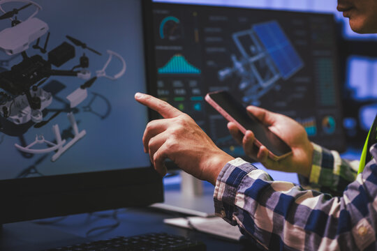 Person pointing at a screen while using a smartphone beside drone control data on a computer monitor in a modern tech workspace.