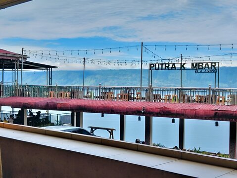 A beautiful view of Putra K Saba Dining, a relaxing dining spot overlooking a stunning natural landscape. The open-air terrace features colorful chairs, a red roof, and decorative string lights.