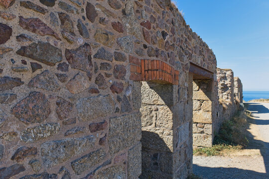 Textured stone wall of ancient ruins features an arched opening, revealing a glimpse of the sea and sky on a bright day - Jacob's Mine, Botallack, Cornwall, UK