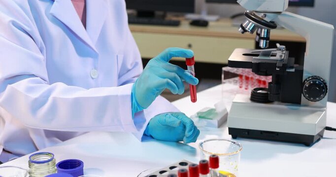Laboratory technician in a white coat and blue gloves holding a blood test tube beside a microscope in a medical lab