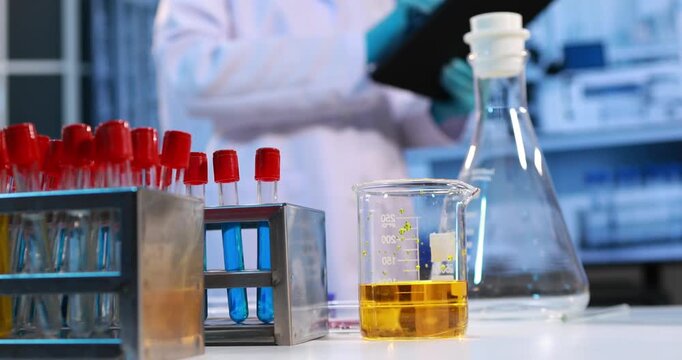 Laboratory scene with test tubes, a glass beaker of yellow liquid, and a flask on a bench, with a scientist blurred in the background, research, testing, and science work.