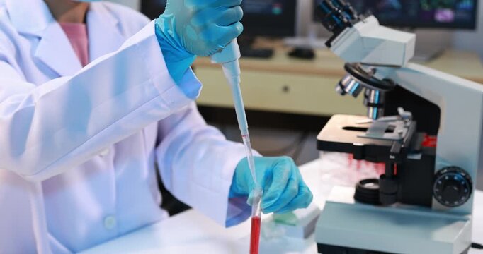 Scientist in a lab coat and blue gloves using a pipette to transfer red liquid into a test tube beside a microscope in a modern laboratory.