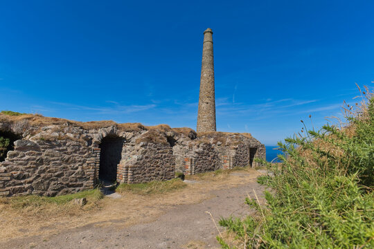 A historic stone building with arched doorways and a tall chimney stands on a grassy cliff under a clear blue sky - Jacob's Mine, Botallack, Cornwall, UK