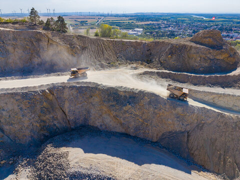 Machines work in a quarry with dust flying around and a view of the landscape far away in the background during daytime