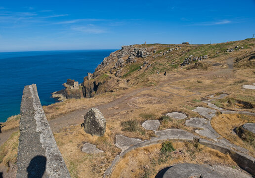 A sunny coastal landscape features ancient concrete ruins on a dry, grassy cliff overlooking a deep blue sea - Jacob's Mine, Botallack, Cornwall, UK