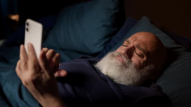 Bearded middle-aged man reads hurtful texts on phone in bed, feeling depressed