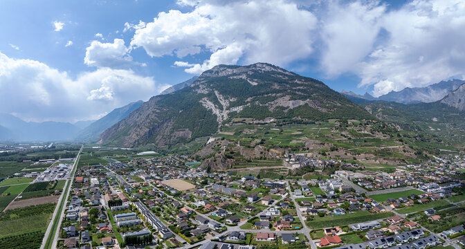 Aerial view of the town of Riddes in the Rhone Valley, featuring the River Rhone, the A9 highway, industrial zones, and surrounding vineyards under a cloudy sky in Riddes, Valais, Switzerland.