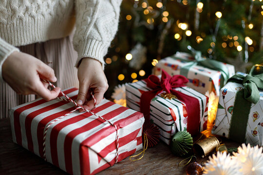 Woman wrapping Christmas present with string at wooden table, hands tying ribbon on striped gift box with festive lights in moody room. festive seasonal preparation
