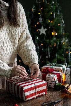 Woman wrapping Christmas present with string at wooden table, hands tying ribbon on striped gift box with festive lights in moody room. festive seasonal preparation