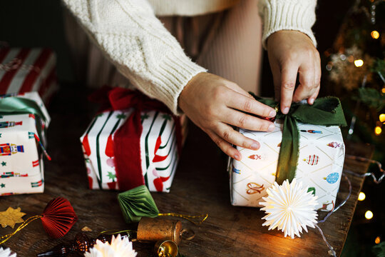 Woman in Cozy Sweater Tying abow Around Wrapped Christmas Present on a Rustic Table with Star Lights and ornaments in festive room. scandinavian holiday decor,  winter celebration