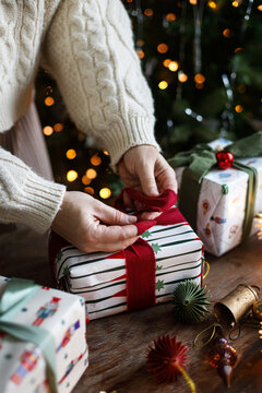 Woman in Cozy Sweater Tying a Red bow Around Wrapped Christmas Present on a Rustic Table with Star Lights and ornaments in festive room. scandinavian holiday decor,  winter celebration