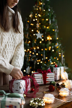 Woman in cozy sweater wrapping gift boxes on wooden table near decorated glowing Christmas tree in moody room. stylish gift ideas, Scandinavian holiday decor, and winter celebration