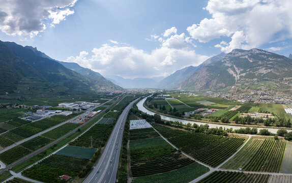 Aerial view of the A9 highway, River Rhone, and industrial zone surrounded by agricultural fields and mountains in Riddes, Valais, Switzerland.