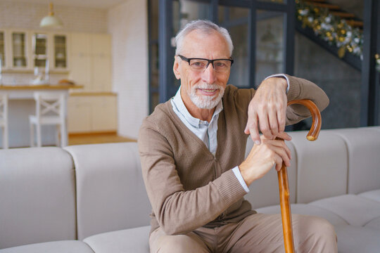 Happy elderly man in eyeglasses holding wooden cane and sitting on couch at home, smiling senior male indoors, positive emotion, retirement lifestyle and mobility support healthcare concept