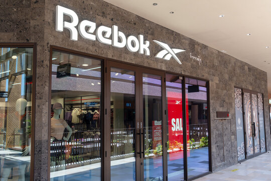Sira village mall, Bali, Indonesia, August 16,2026: Illuminated Reebok storefront featuring the brand logo, textured volcanic stone facade wall, double glass doors, mannequin displays, and sale signs 