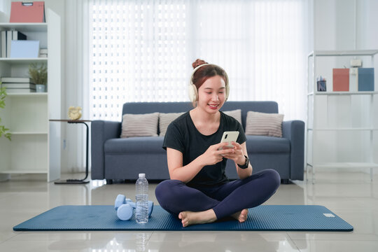 Young Asian woman using smartphone to follow online fitness class while sitting on yoga mat at home with copy space