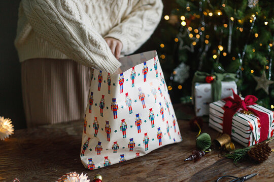Woman in knitted sweater wrapping Christmas gift box with nutcracker pattern paper on table, concept of holiday preparation process, DIY craft gift wrapping, and cozy winter lifestyle