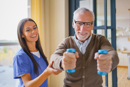 Elderly man doing dumbbell exercises with help of young female nurse at home, senior patient receiving rehabilitation support, healthcare assistance and home care concept