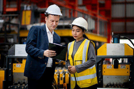 Two professional engineer,worker,technician use clipboard discuss work, walk in steel metal manufacture factory plant industry. Black African American man and woman wear hard hat check quality machine
