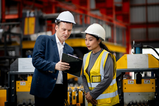 Two professional engineer,worker,technician use clipboard discuss work, walk in steel metal manufacture factory plant industry. Black African American man and woman wear hard hat check quality machine