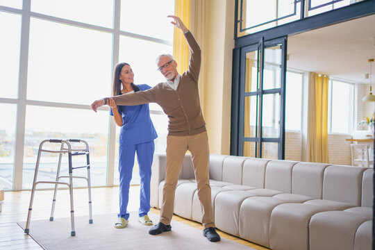 Young female nurse helping senior man stretch his arm at home, caregiver supporting elderly patient during rehabilitation exercise, mobility recovery, home care service and healthcare concept