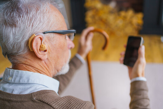 Back view of senior man with walking cane holding mobile phone with black blank mockup screen, elderly male at home using smartphone, copy space, modern technology and retirement lifestyle concept