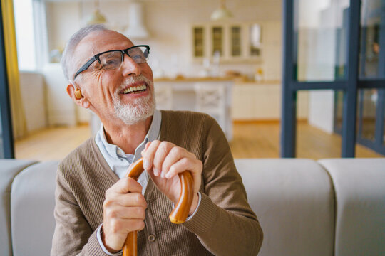 Happy senior man with wooden cane and hearing aid sitting on couch and laughing at home, elderly male enjoying comfortable retirement lifestyle, positive emotion and indoor portrait concept