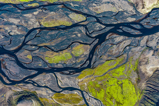 Aerial view of a braided river system with dark volcanic sediment and vibrant green mossy patches flowing through the landscape of Iceland.