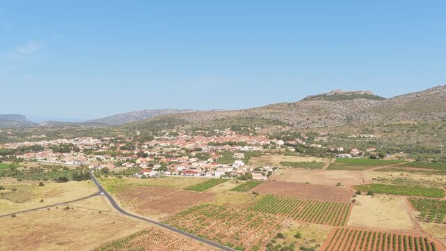 The rural village of Opoul Perillos in southern France. The video shows the village surrounded by agricultural fields, vineyards, and rolling hills under a clear blue sky, aerial view.